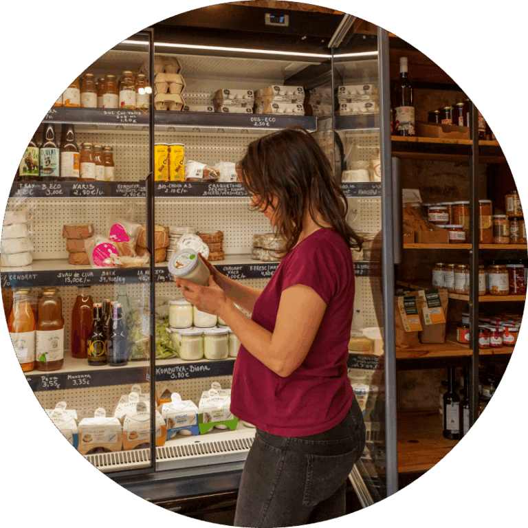 Una dona davant d'una nevera de productes refrigerats mirant l'etiqueta. Al fons hi ha altres prestatges amb productes. És dins d'una botiga de queviures.